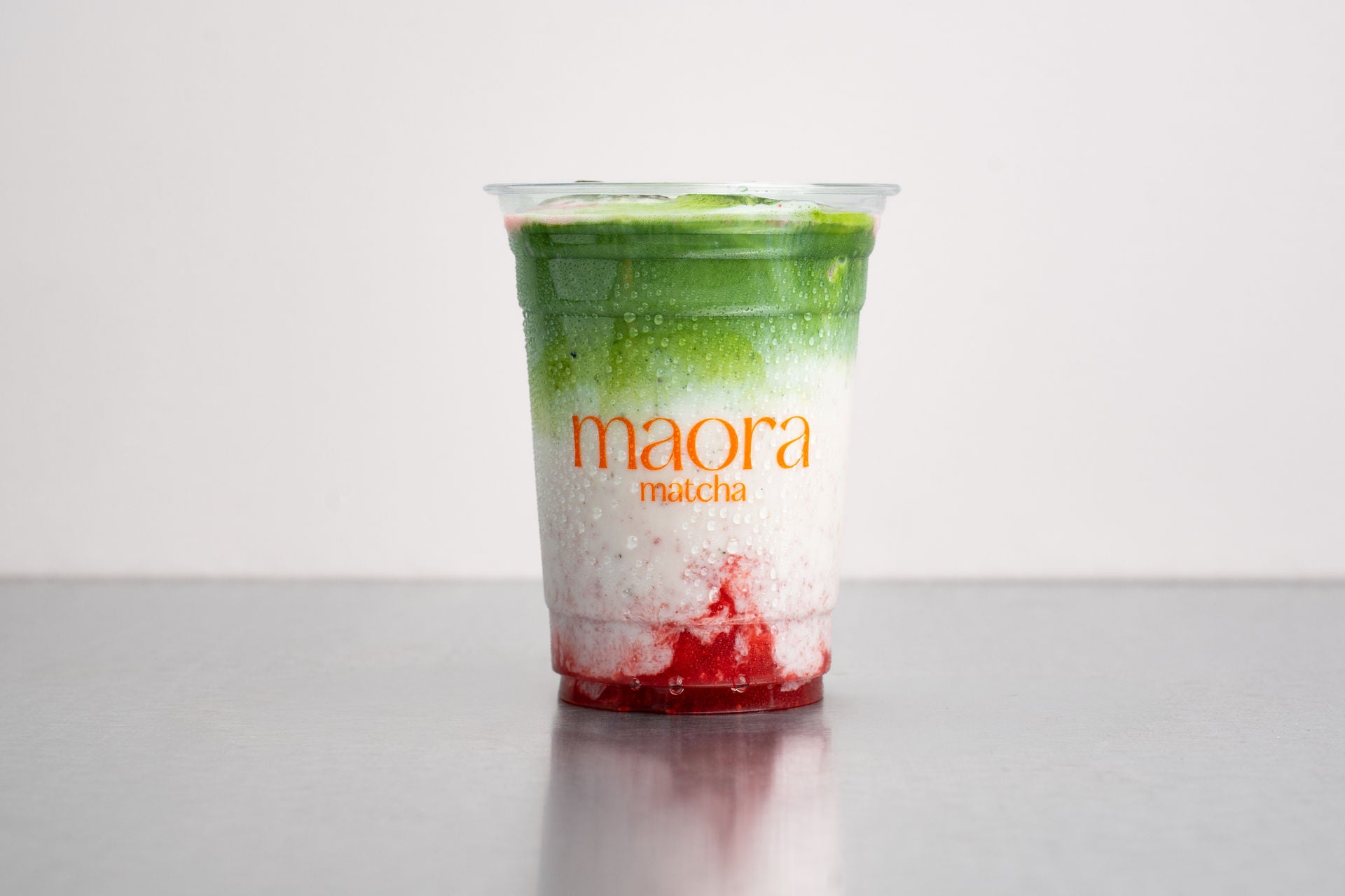 Geschichteter Matcha Drink mit Erdbeerschicht im Becher mit Maora Logo