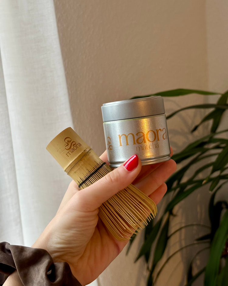 Maora Matcha Dose und Bambus Matcha Besen (Chasen) in einer Hand vor neutralem Hintergrund