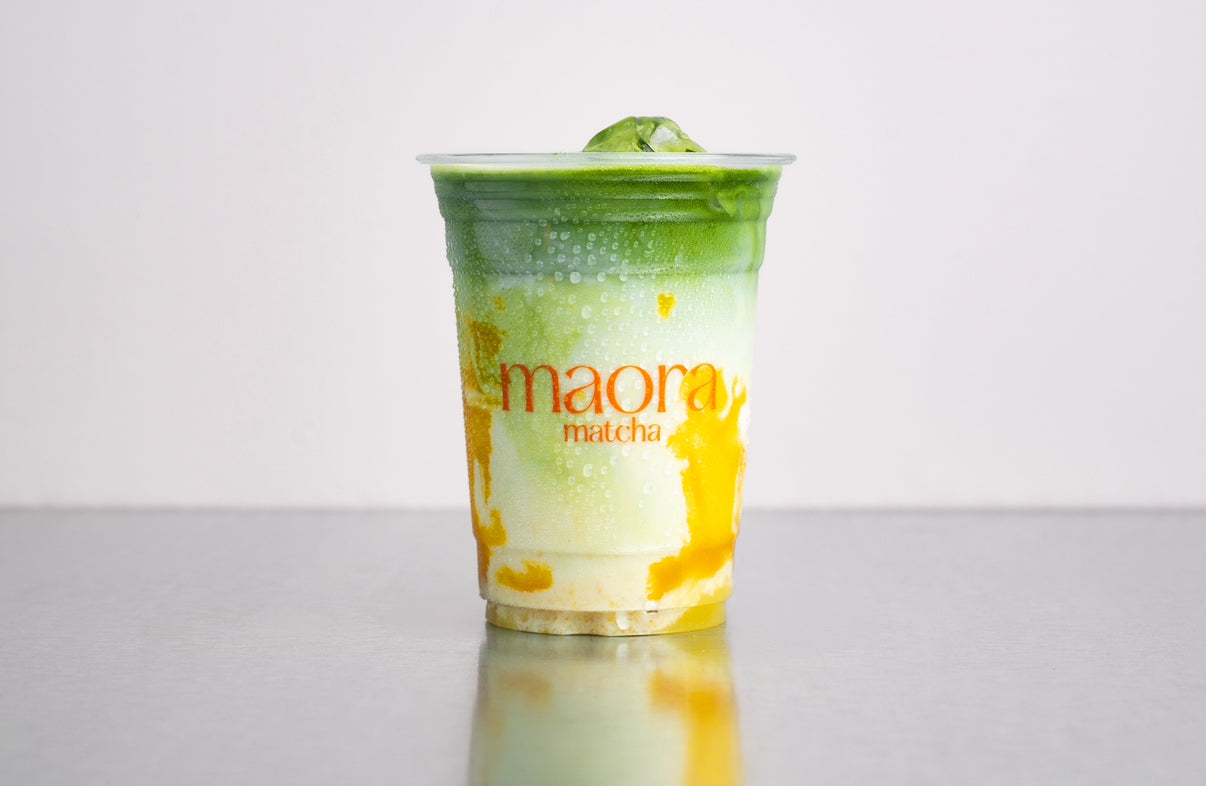 Geschichteter Matcha Drink mit Mango Frucht Püree im Becher mit Maora Logo
